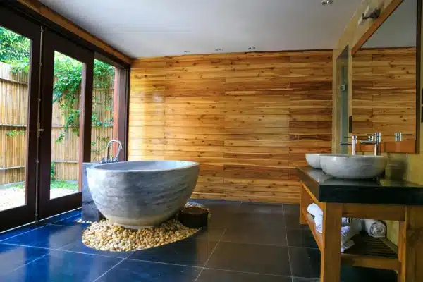 Spa-style bathroom featuring a stone bathtub, wood-panelled wall, dark flooring, vessel sinks and a large glass window overlooking the garden.