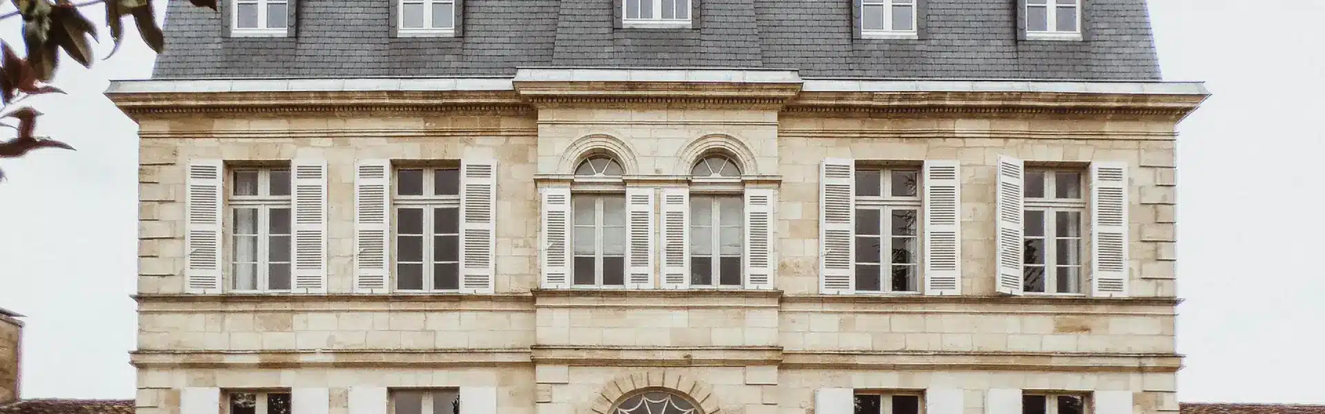 French manor house façade in cut stone with slate roof, dormer windows, white shutters and arched front entrance.